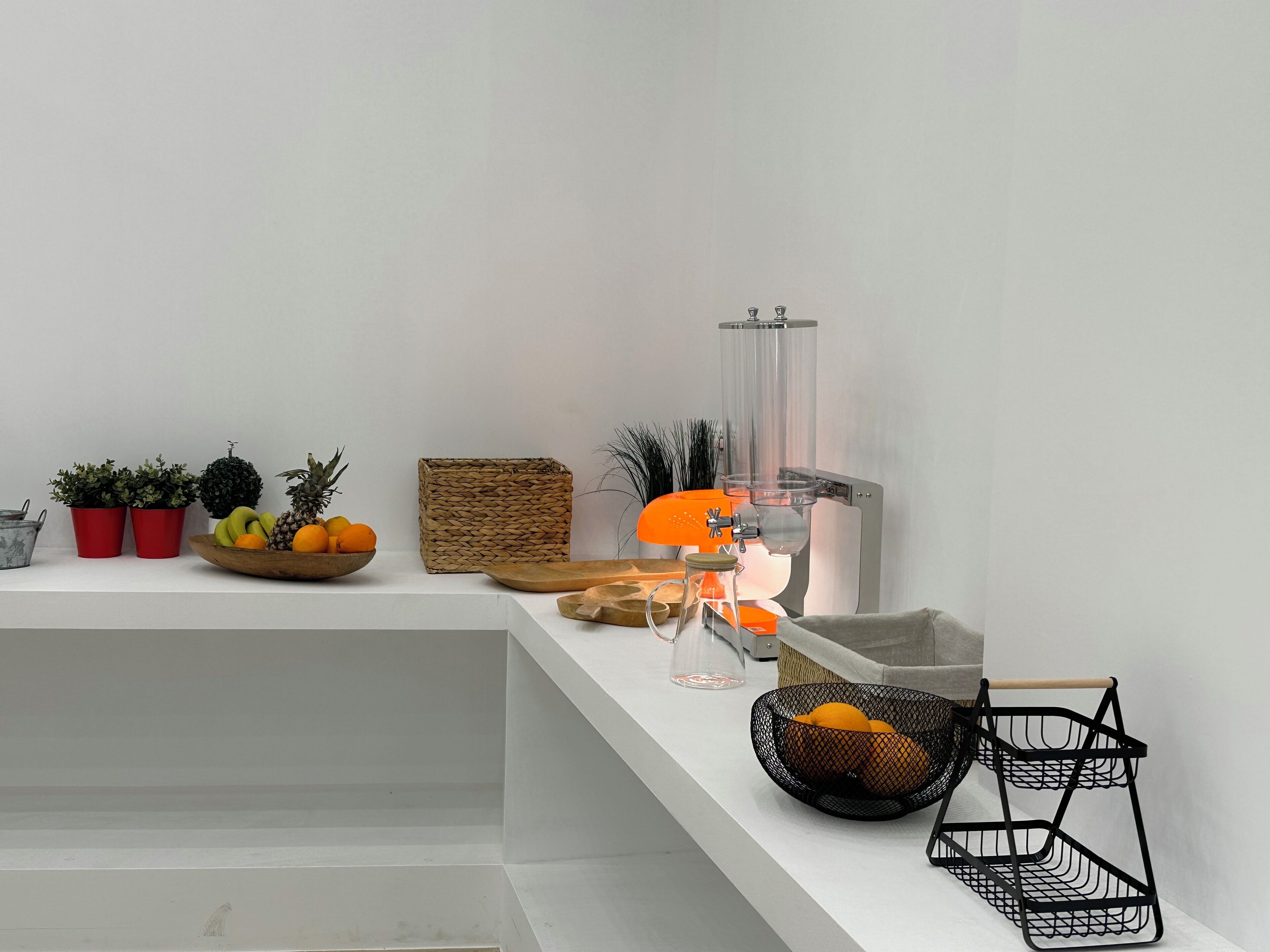 A modern, minimalist room with a white wall, a wooden bench, a fruit bowl, potted plants, a woven basket, a glass pitcher, and a black wire basket.