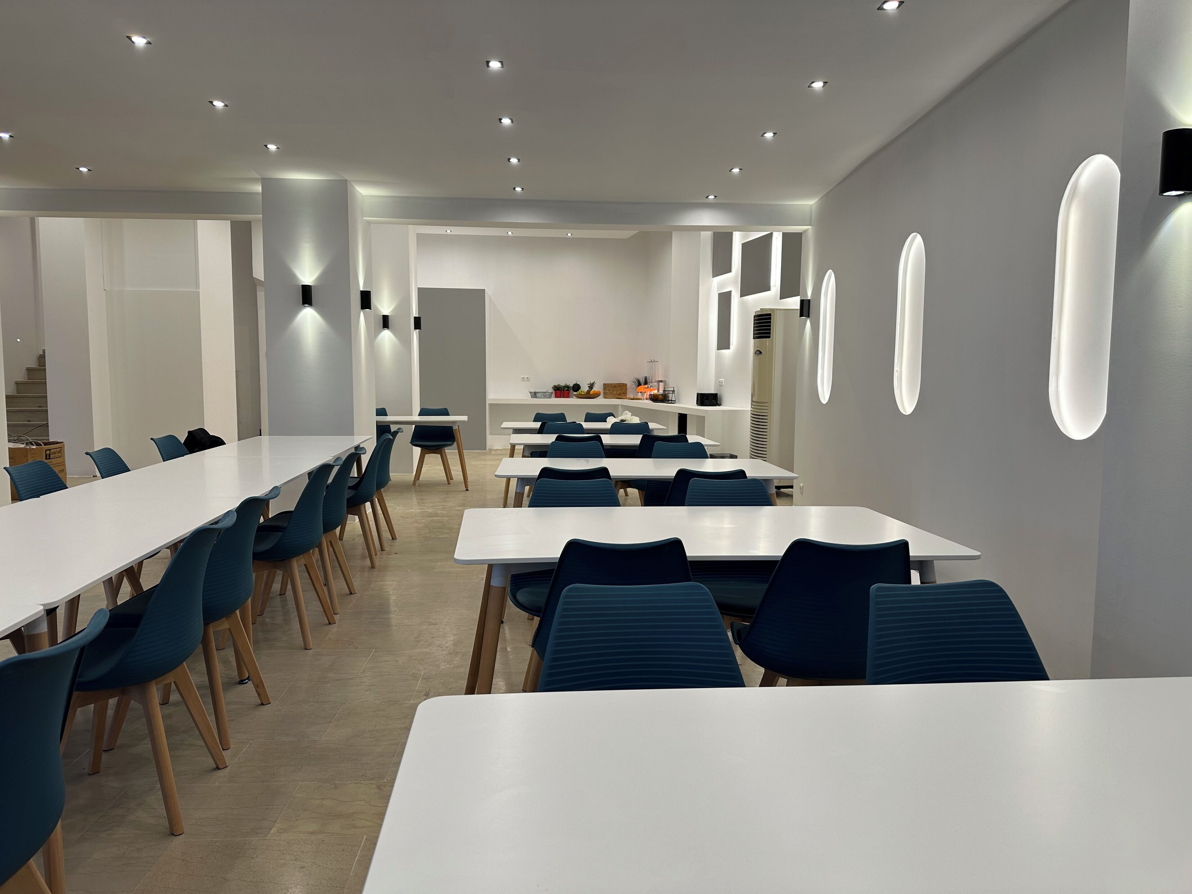 Un comedor moderno con mesas blancas y sillas azules, luces empotradas en el techo e iluminación adosada a las paredes.