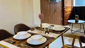 Un comedor con una mesa larga dispuesta para cuatro, con platos, fuentes y vasos blancos. Hay un pequeño jarrón con flores sobre la mesa.
