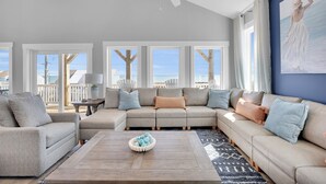 Ein geräumiges Wohnzimmer mit einem großen Ecksofa, einem Couchtisch mit Dekoration und Blick ins Freie durch große Fenster.