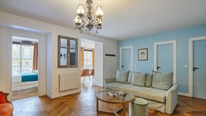 Un salón con piso de madera en patrón de espiga, un sofá color beige, una mesa de centro de vidrio y una araña. Se observa una puerta con un marco en la pared.