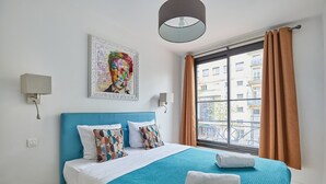 Un dormitorio con una cama grande, una ventana con cortinas, una lámpara de pared y un cuadro colorido.