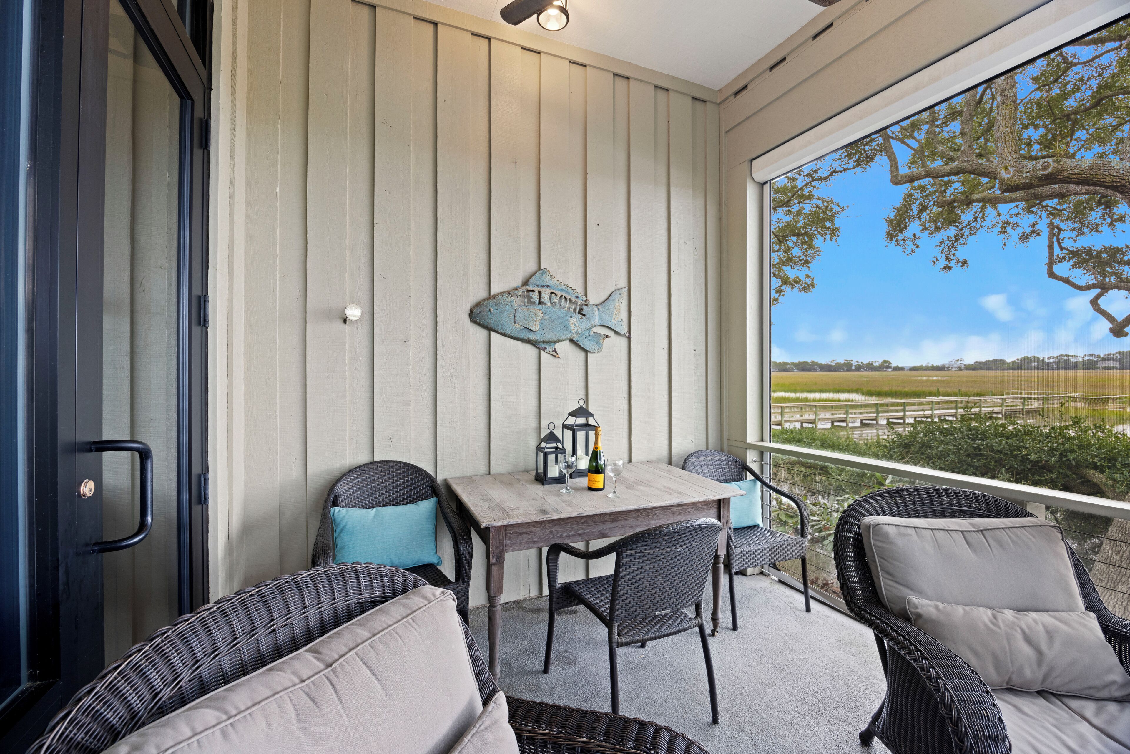 A covered patio with wicker furniture, a table with a bottle and glasses, and a wall-mounted fish sign.