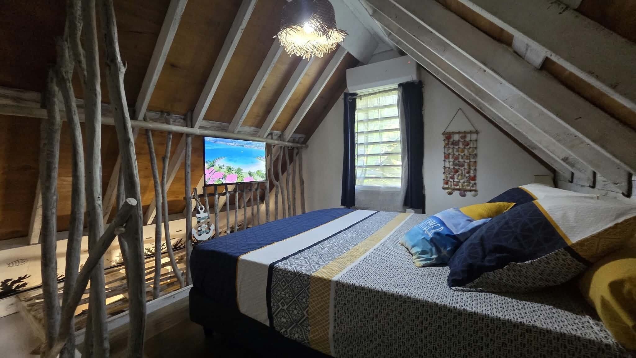 A loft bedroom with a sloped ceiling, a bed with patterned bedding, a television on the wall, and a window with curtains.