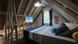 A loft bedroom with a sloped ceiling, a bed with patterned bedding, a television on the wall, and a window with curtains.