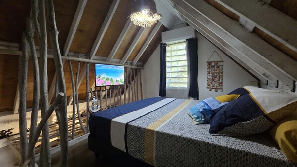 A loft bedroom with a sloped ceiling, a bed with patterned bedding, a television on the wall, and a window with curtains.