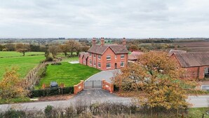 Een groot, bakstenen huis met een hekwerk ervoor, omringd door groene velden en bomen.