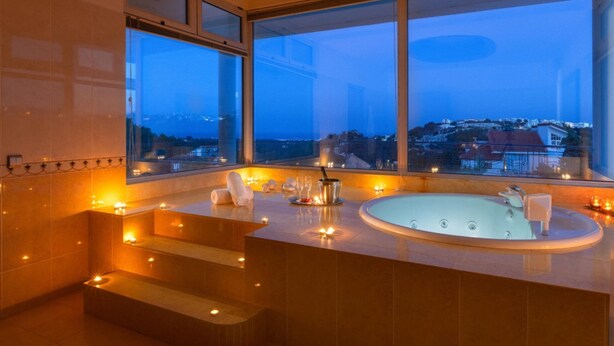 Una spa moderna con una vasca da bagno ampia, candele e vista sulla città al crepuscolo.