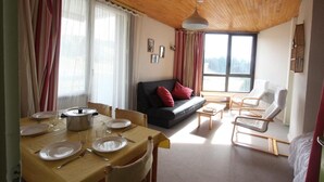 Una sala de estar acogedora con techo de madera, un comedor con mesa dispuesta para dos personas, un sofá con cojines rojos y un amplio ventanal con vistas al exterior.