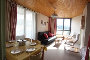 Una sala de estar acogedora con techo de madera, un comedor con mesa dispuesta para dos personas, un sofá con cojines rojos y un amplio ventanal con vistas al exterior.