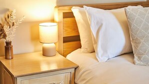 A neatly made bed with white linens and a striped pillow, a bedside lamp, and a vase with dried plants on a wooden nightstand.