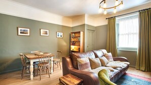 A cozy living room with a leather sofa, wooden dining table, and chairs. A bookshelf and a door are visible in the background.