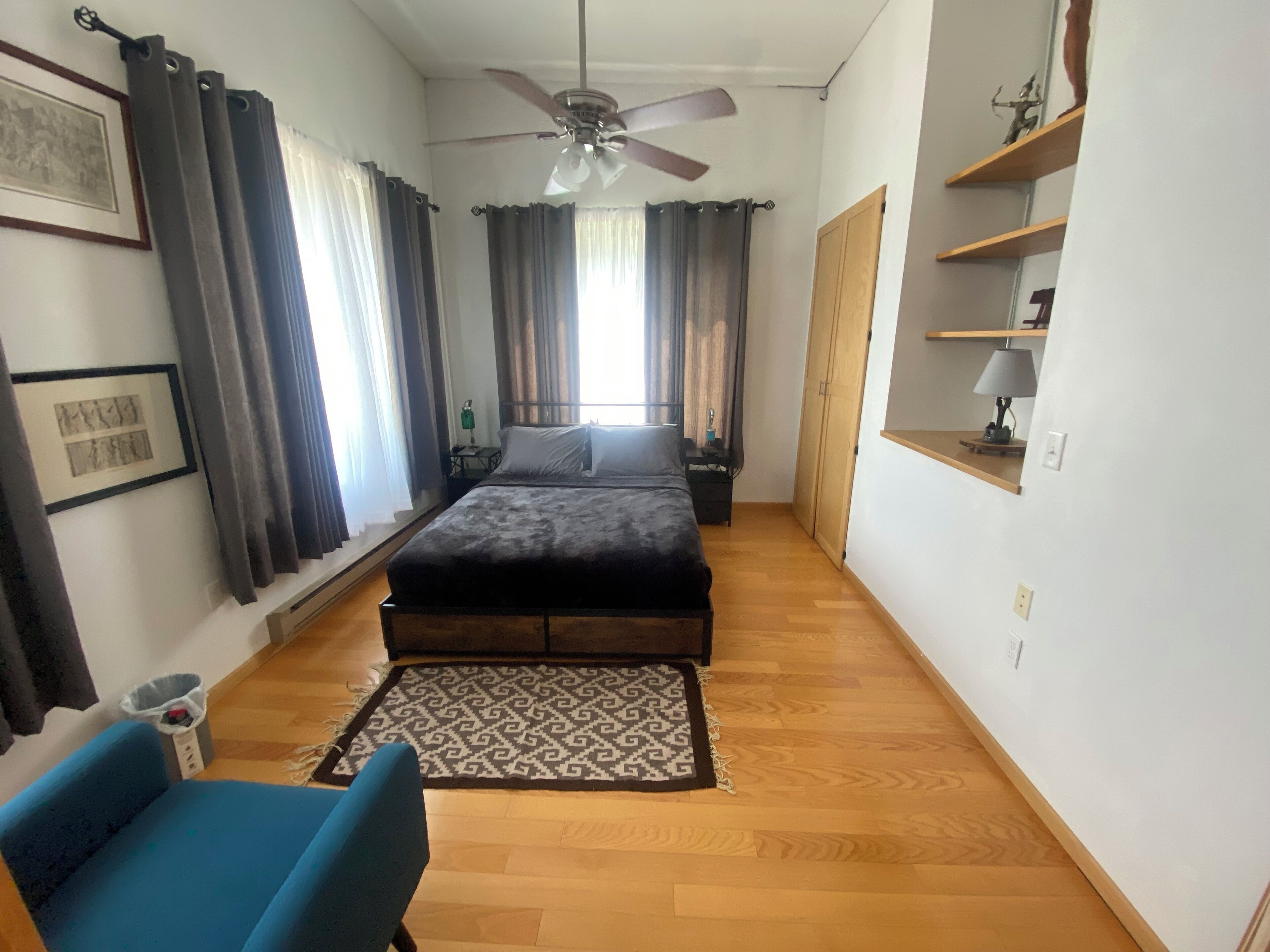 A bedroom with a bed, a blue chair, a ceiling fan, a wooden floor, and a patterned rug.
