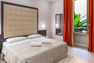 Habitación de hotel con una cama grande, cabecero, mesita de noche con lámpara, espejo y ventana con cortinas.