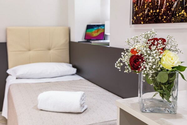 A neatly made bed with a white pillow and a rolled white towel, a clear glass vase with red and yellow roses, and a laptop on a desk in the background.