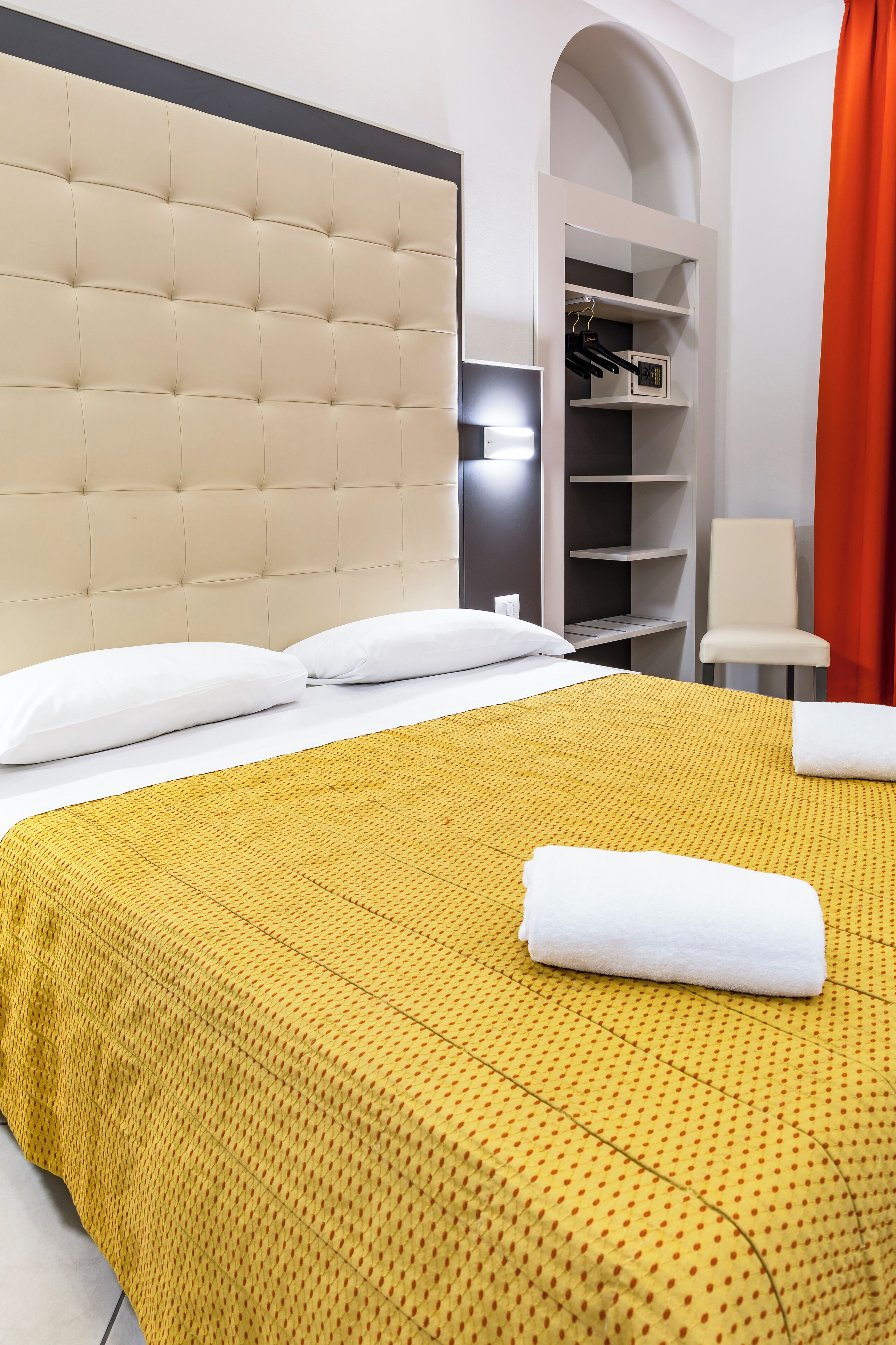 Una habitación de hotel con cabecera capitonada, colcha amarilla estampada, almohadas blancas, toalla blanca, armario con estantes y una cortina roja.