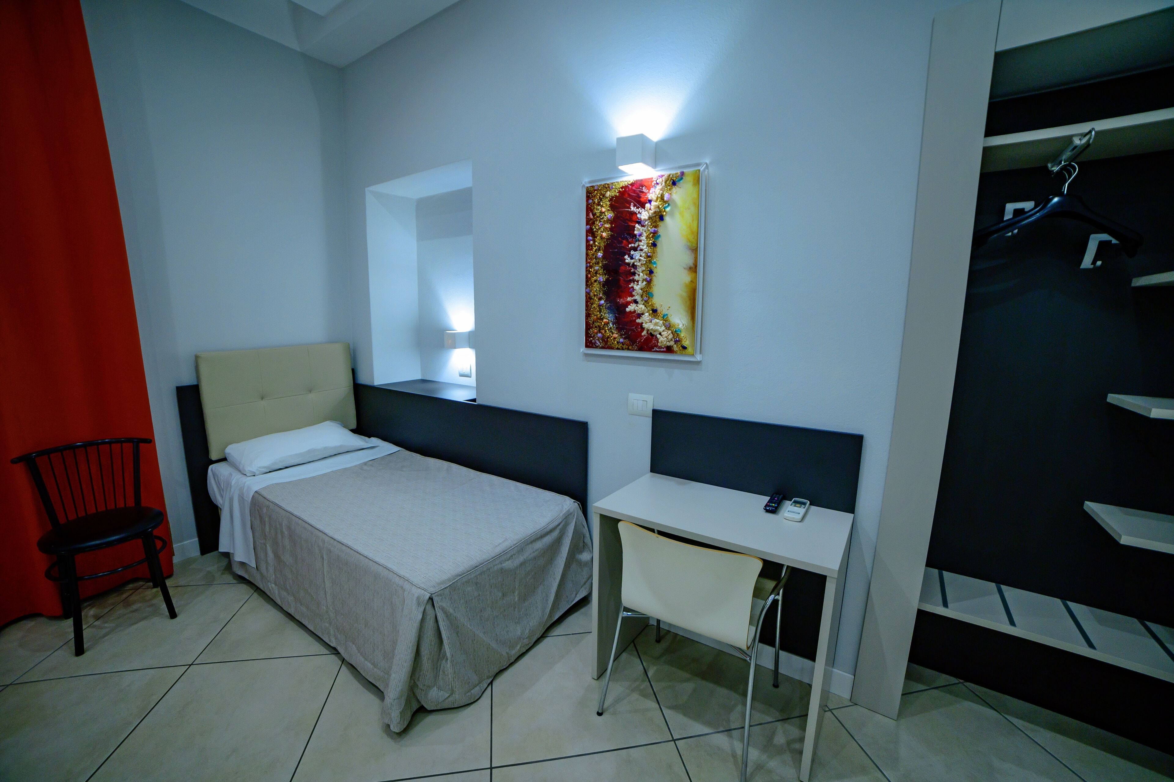 Ein Hotelzimmer mit Bett, Schreibtisch, Stuhl, einem roten Vorhang und einem Wandbild.