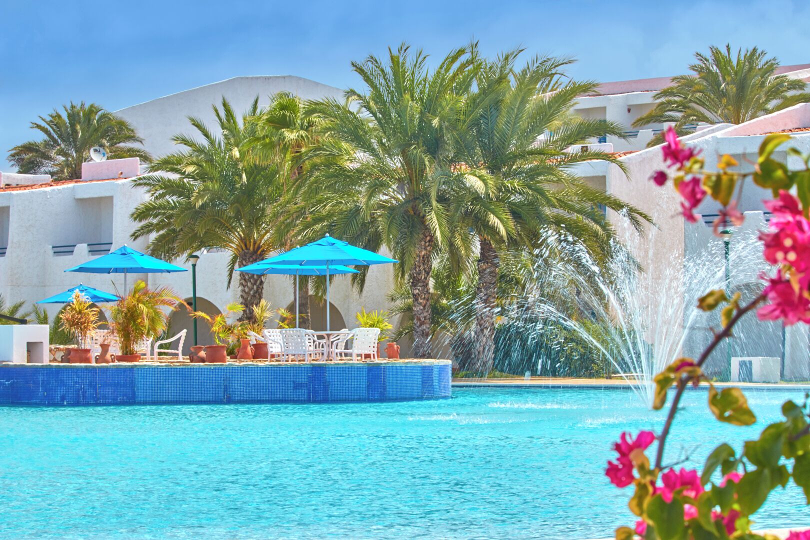 Piscina con fuente de agua, rodeada de palmeras y sombrillas azules.
