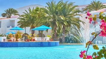 Piscina con fuente de agua, rodeada de palmeras y sombrillas azules.