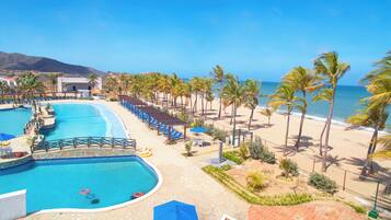 A resort with a large swimming pool, beach area with palm trees, and a mountain in the background.