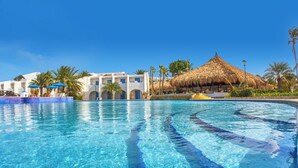 Une piscine d’eau cristalline, avec une structure à toit de chaume en arrière-plan, entourée de palmiers et d’un bâtiment blanc agrémenté de touches de bleu.