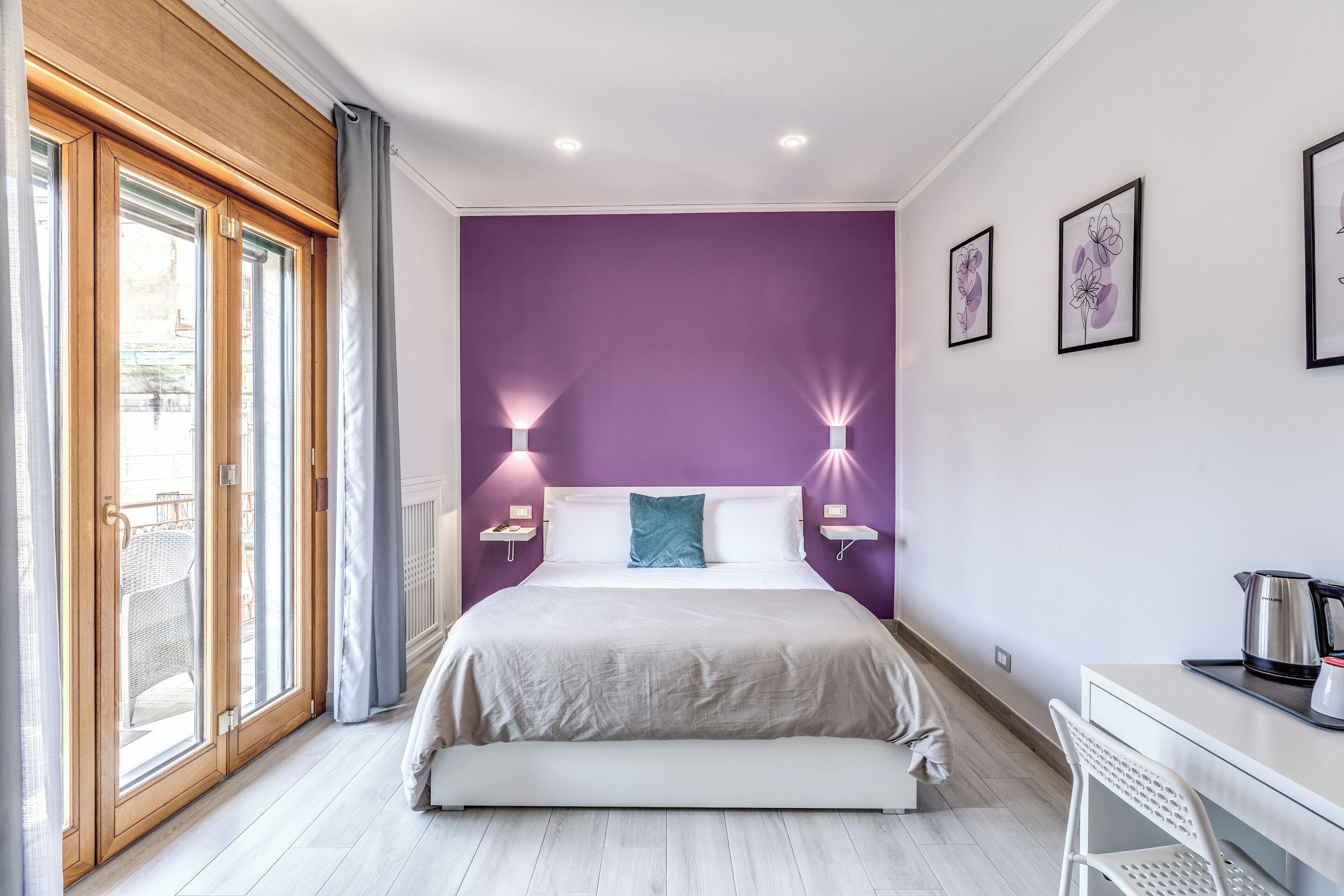 Ein Schlafzimmer mit einem Bett, zwei Nachttischlampen, einem Schreibtisch mit Wasserkocher und einem Fenster mit Vorhängen.