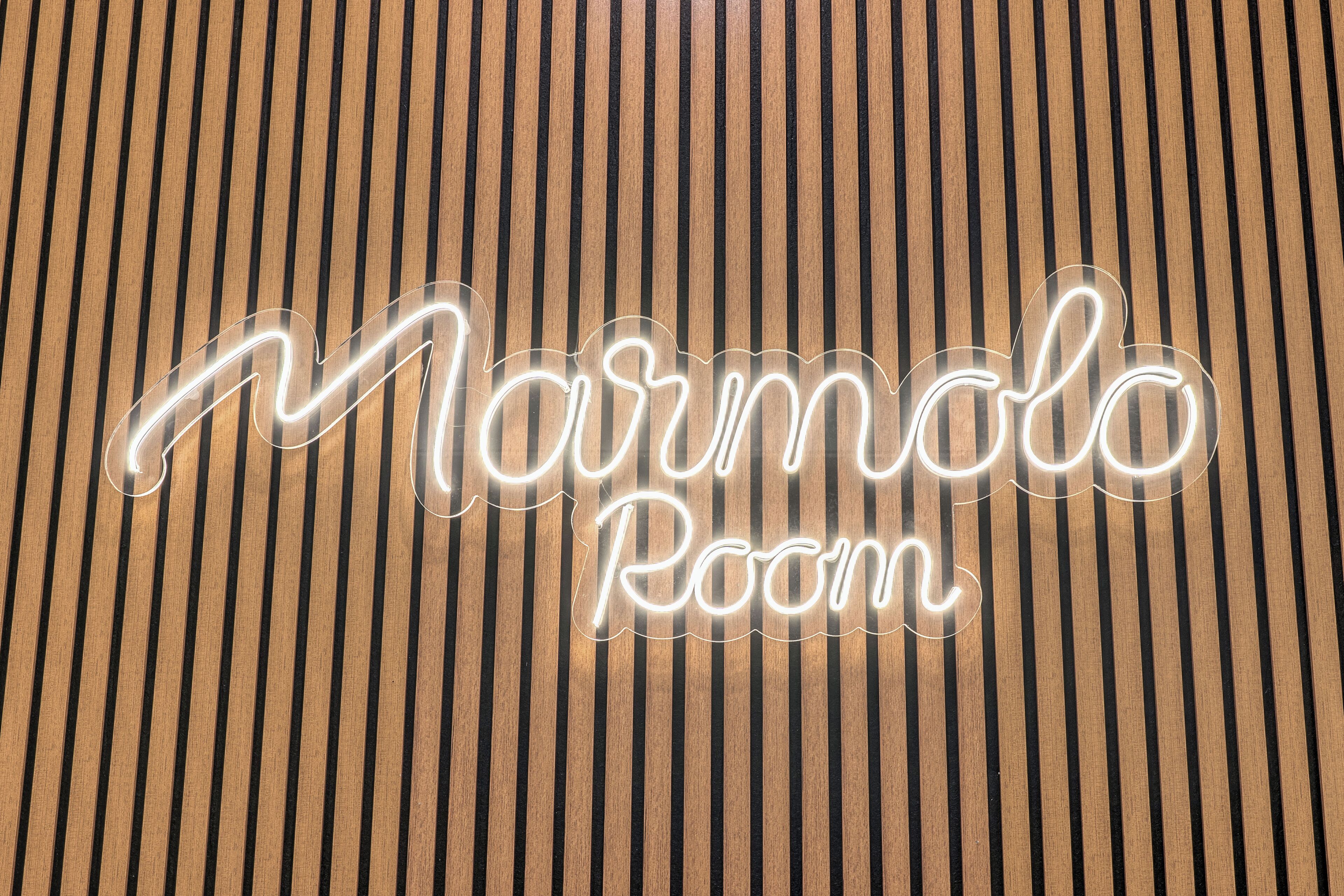 Een neonbord waarop 'Marmolo Room' staat, op een houten oppervlak met verticale lijnen.