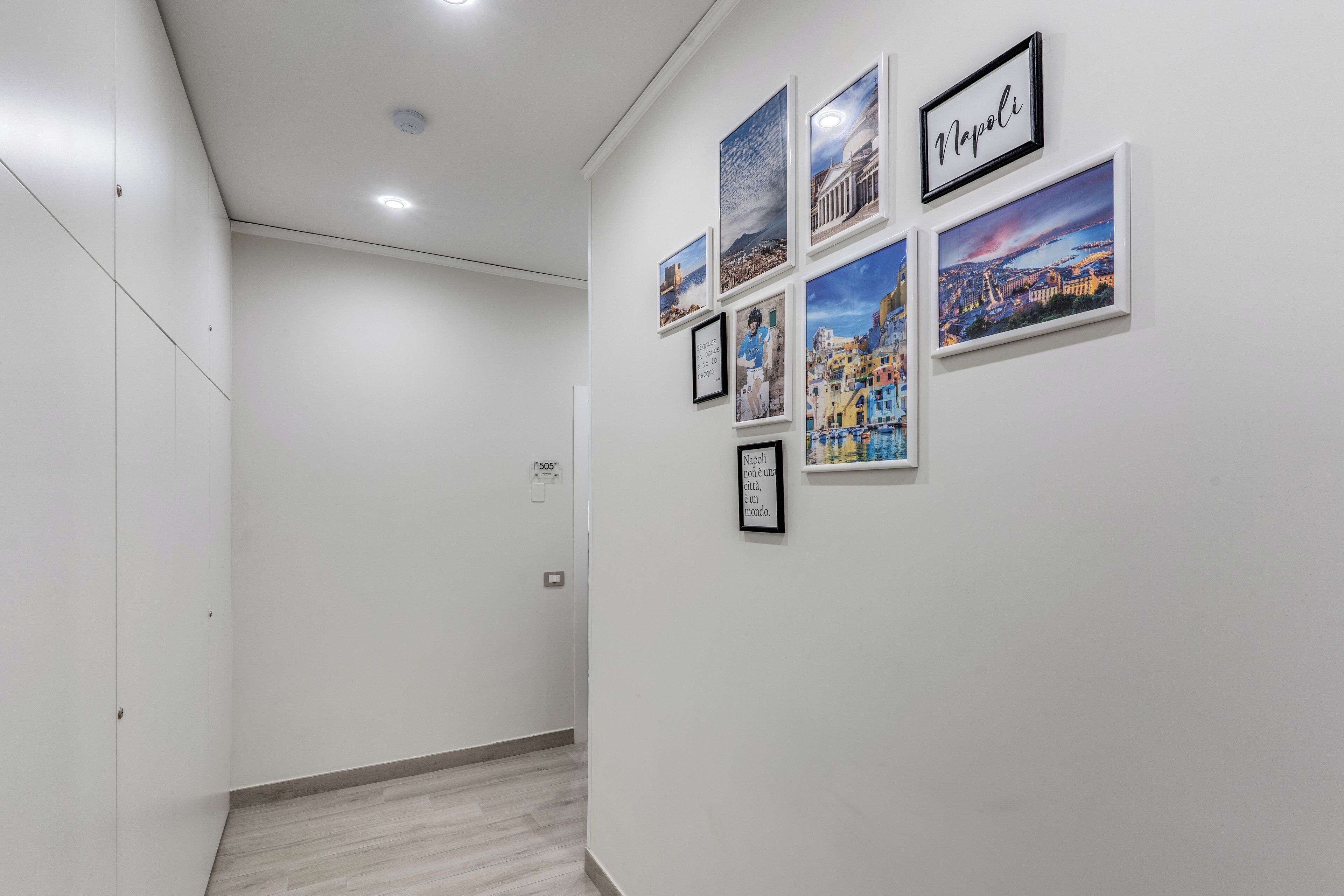 A hallway with white walls and a wooden floor. There are framed pictures on the wall, including one labeled "Napoli".