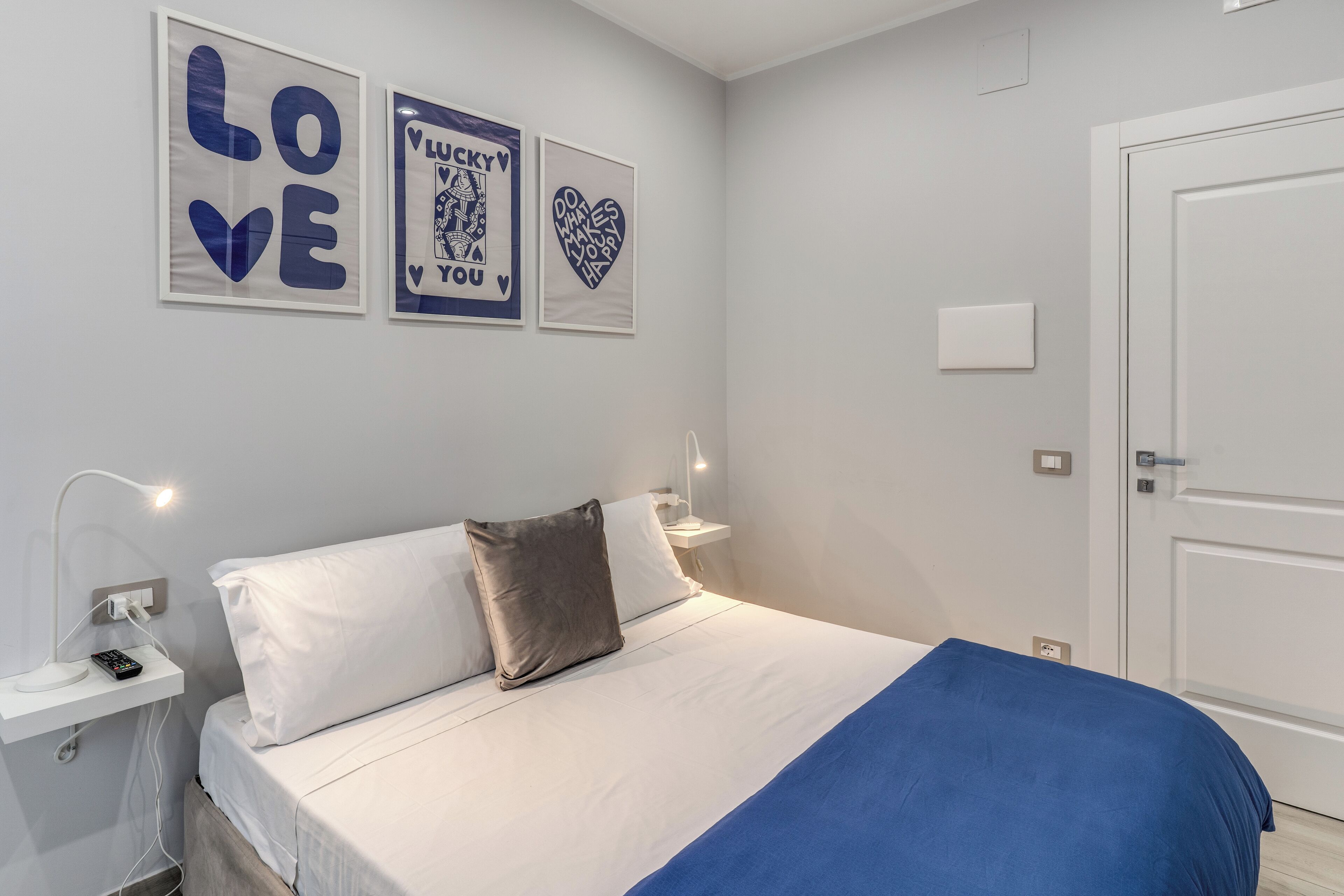 A neatly made bed with white and blue bedding, a bedside table with a lamp, and three framed wall art pieces with text and heart designs.