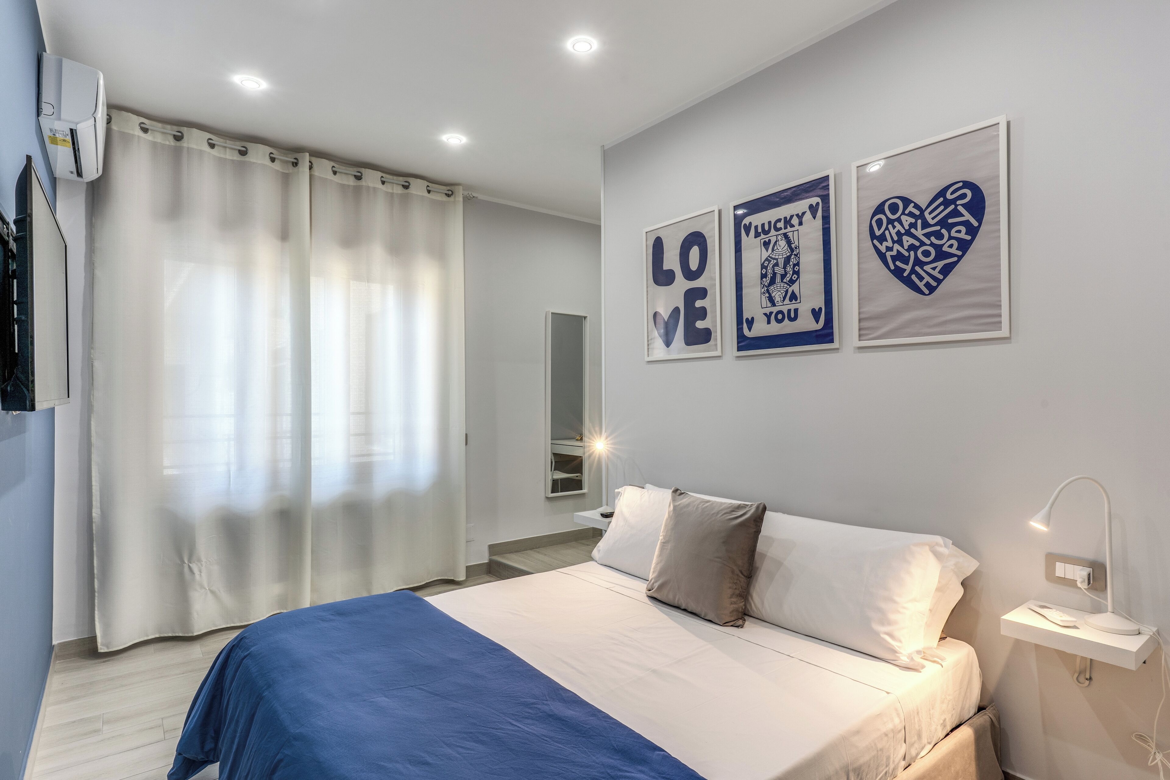 A bedroom with a bed, white curtains, a bedside table with a lamp, and wall art.