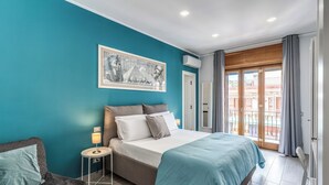 A bedroom with a bed, a nightstand, a chair, a window with a view of buildings, and a wall with a framed picture. - Marmolo Room (Naples)
