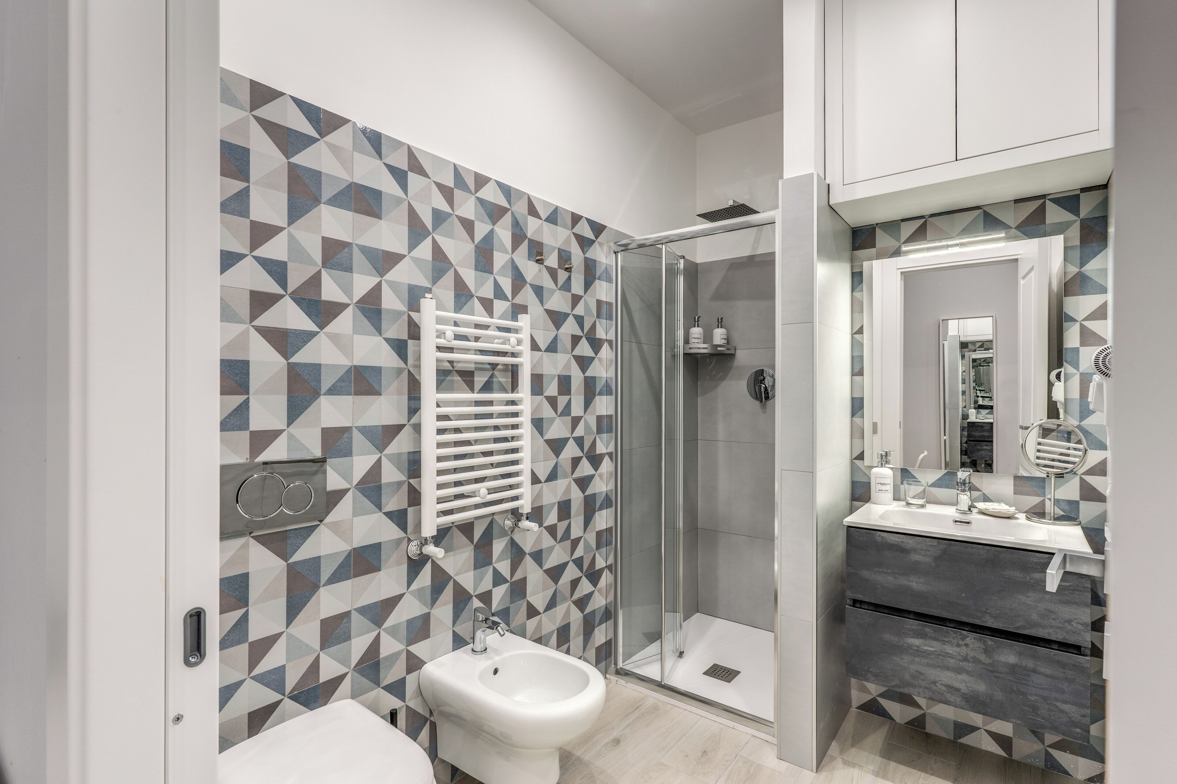 A modern bathroom with a geometric tile backsplash, a glass shower enclosure, a bidet, and a vanity with a mirror.