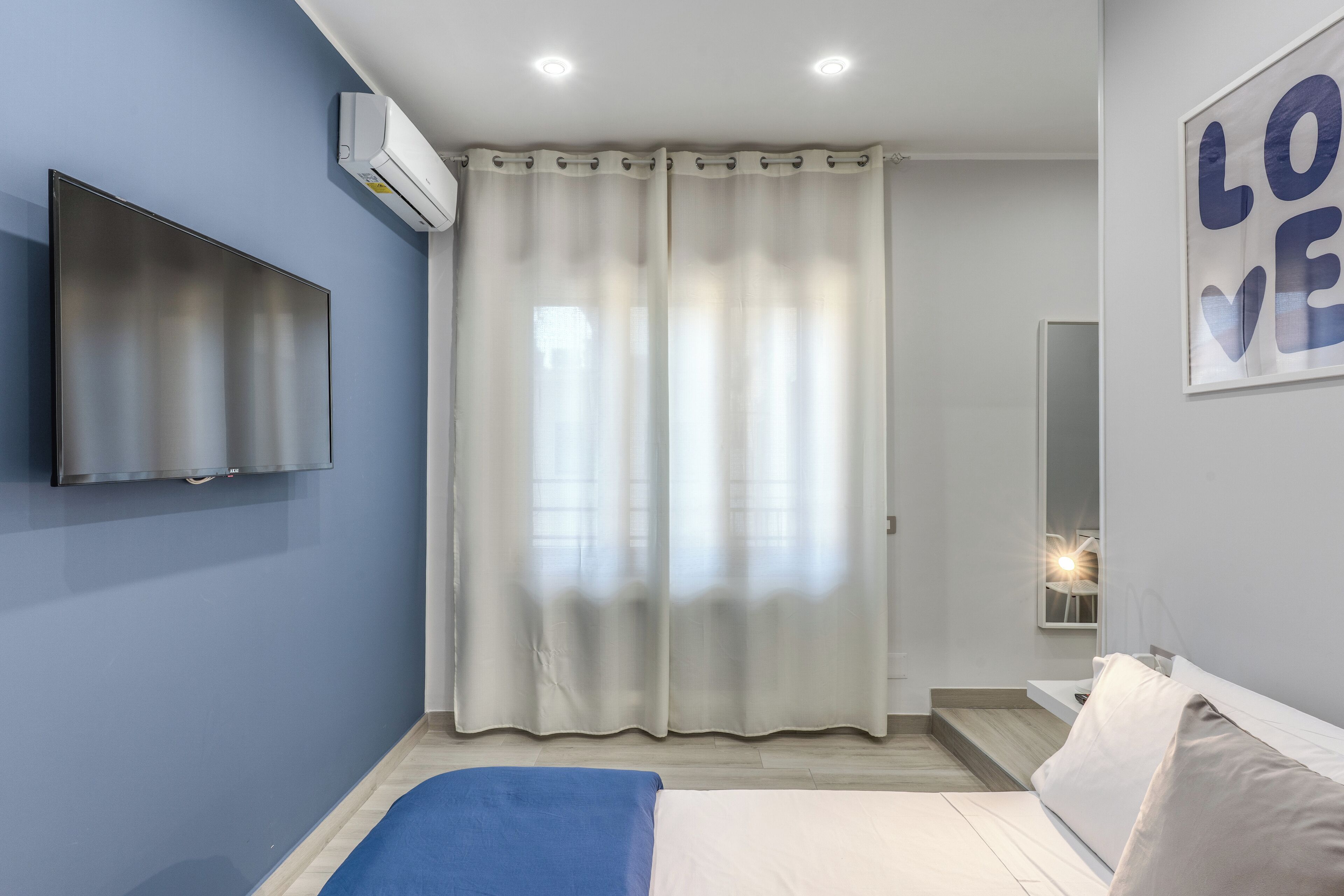 A bedroom with a bed, a flat-screen TV mounted on the wall, a large window with sheer curtains, and a wall-mounted mirror.
