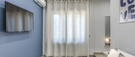 A bedroom with a bed, a flat-screen TV mounted on the wall, a large window with sheer curtains, and a wall-mounted mirror.