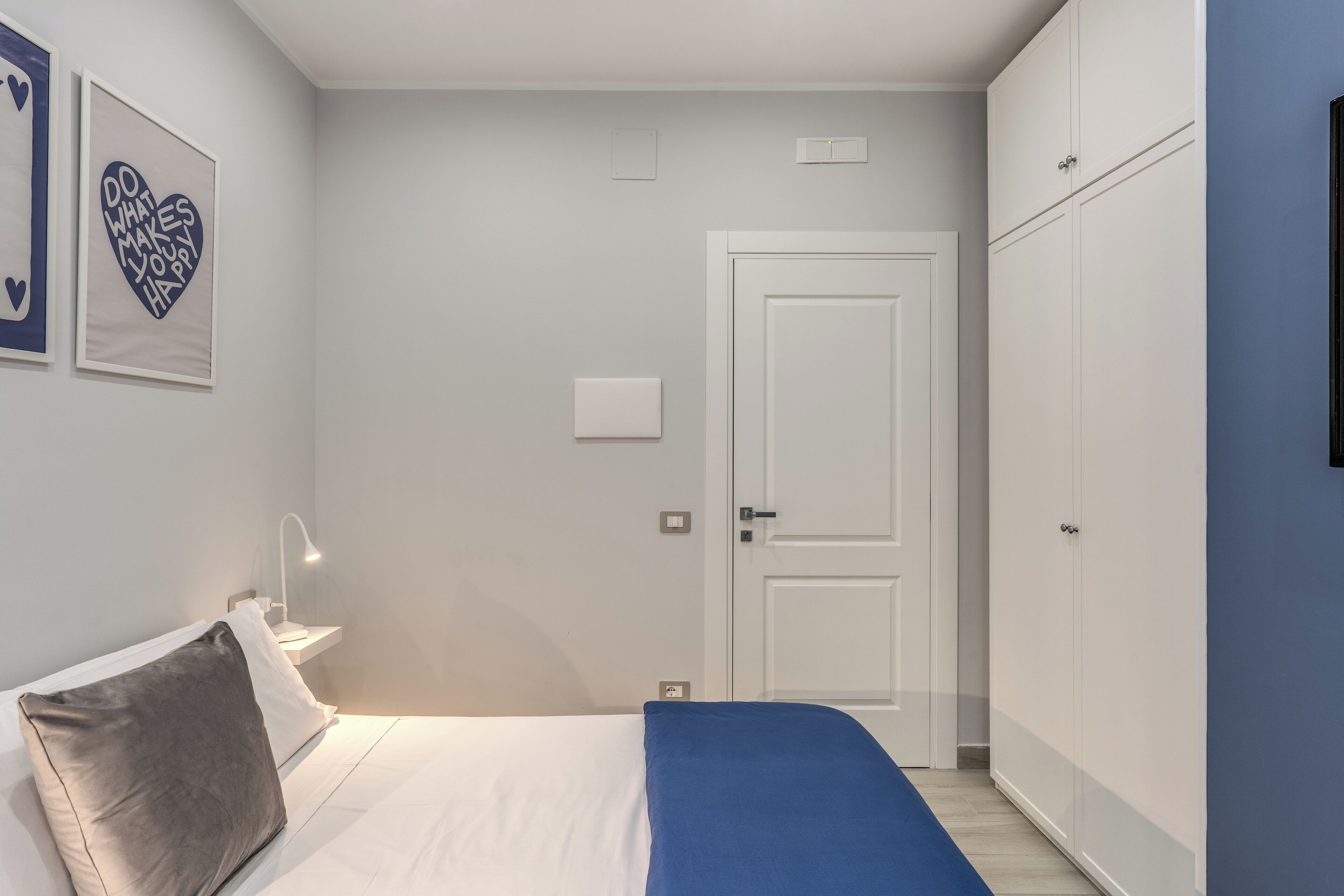 A bedroom with a bed, a nightstand, a desk lamp, a wardrobe, and two framed pictures on the wall.