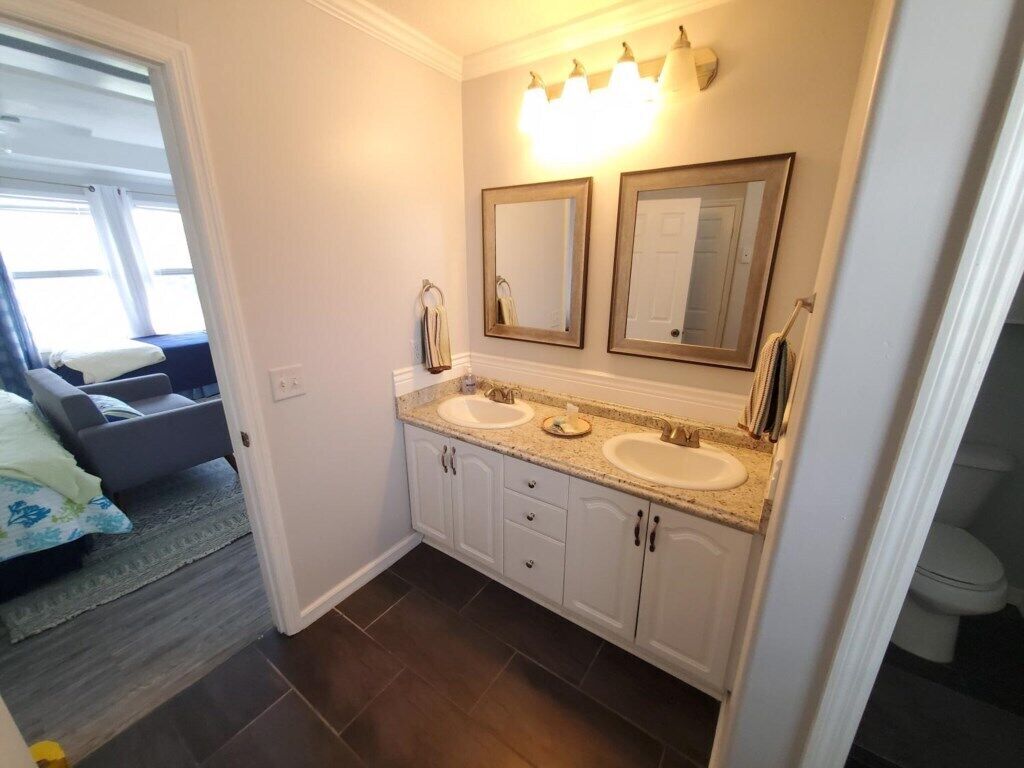 A bathroom with two sinks, a vanity, and a mirror. There is a window with blinds and a view of the outside.