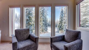 A room with three large windows offering a view of snow-covered trees. There are two armchairs in the room. - Pet friendly chalet right on the ski way, steps from tube town pet friendly too! (Silver Star Mountain)