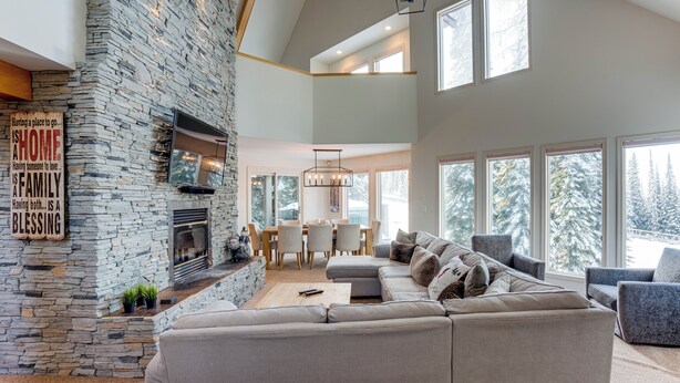 A modern living room with a stone fireplace, a large sectional sofa, and a dining area with a chandelier. - Pet friendly chalet right on the ski way, steps from tube town pet friendly too! (Silver Star Mountain)
