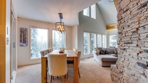 A spacious dining area with a wooden table and chairs, large windows with a snowy view, a stone accent wall, and a modern chandelier. - Pet friendly chalet right on the ski way, steps from tube town pet friendly too! (Silver Star Mountain)