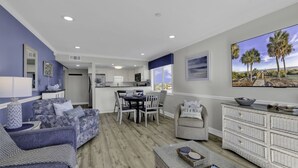 A modern living room with a dining area, a kitchen in the background, and a large window with a view of palm trees. - Bluewater Resort V408A (Myrtle Beach)