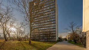 Ein hohes Wohngebäude mit vielen Fenstern und Balkonen, umgeben von Bäumen und einem gepflasterten Gehweg.