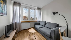 Ein modernes Wohnzimmer mit einem grauen Sofa, einem kleinen runden Tisch, einem Fernseher auf einem Holzgestell und einer Stehlampe.