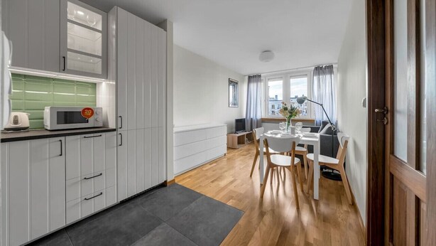 Eine moderne Küche mit grauen Schränken, einer Mikrowelle und einem Wasserkocher. Es gibt einen Essbereich mit Tisch und Stühlen, sowie ein Fenster mit Vorhängen.