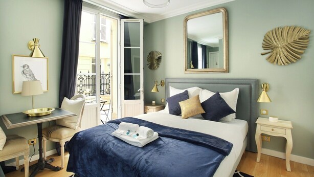 Une chambre d’hôtel comprenant un lit, un bureau, une chaise, une petite table, une table de chevet, un miroir et une fenêtre avec des rideaux.