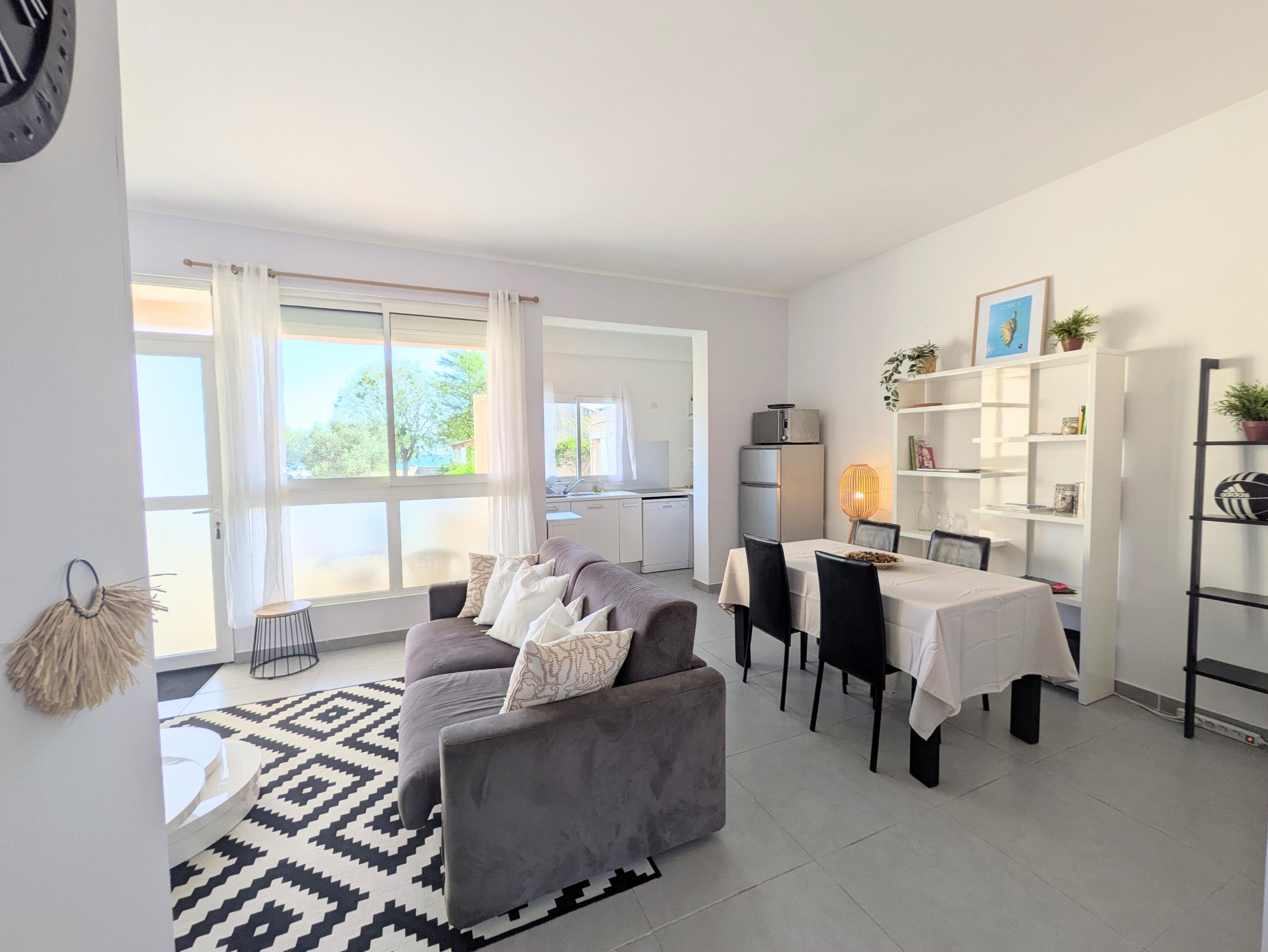 A modern living room with a grey sofa, dining table, and chairs. There is a kitchen in the background, a bookshelf, and a large window with curtains.