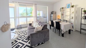 A modern living room with a grey sofa, dining table, and chairs. There is a kitchen in the background, a bookshelf, and a large window with curtains. - La Perla di Santa Maria - Vue mer, plage à 100 m (Santa-Maria-Poggio)