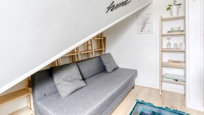 Un canapé gris placé sous un plafond en pente, avec un panneau indiquant « chez soi », des étagères en bois et un tableau encadré accroché au mur.