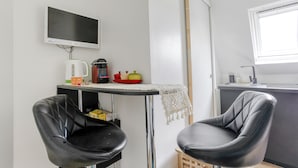 Une petite cuisine équipée d’un téléviseur, d’une bouilloire et d’une machine à café. On y trouve deux chaises noires et une petite table recouverte d’une nappe en dentelle.