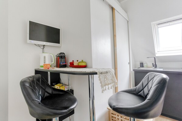 Une petite cuisine équipée d’un téléviseur, d’une bouilloire et d’une machine à café. On y trouve deux chaises noires et une petite table recouverte d’une nappe en dentelle.