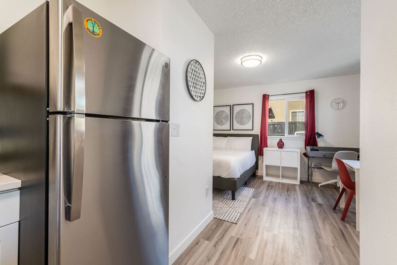 Una cocina moderna con un refrigerador de acero inoxidable, un comedor con una mesa blanca y sillas rojas, y un dormitorio con una cama y una ventana.